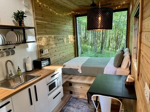 A compact cabin with a kitchenette, a bed with a view of the forest, and a dining area. - The Dogwood Tiny House (Logan)