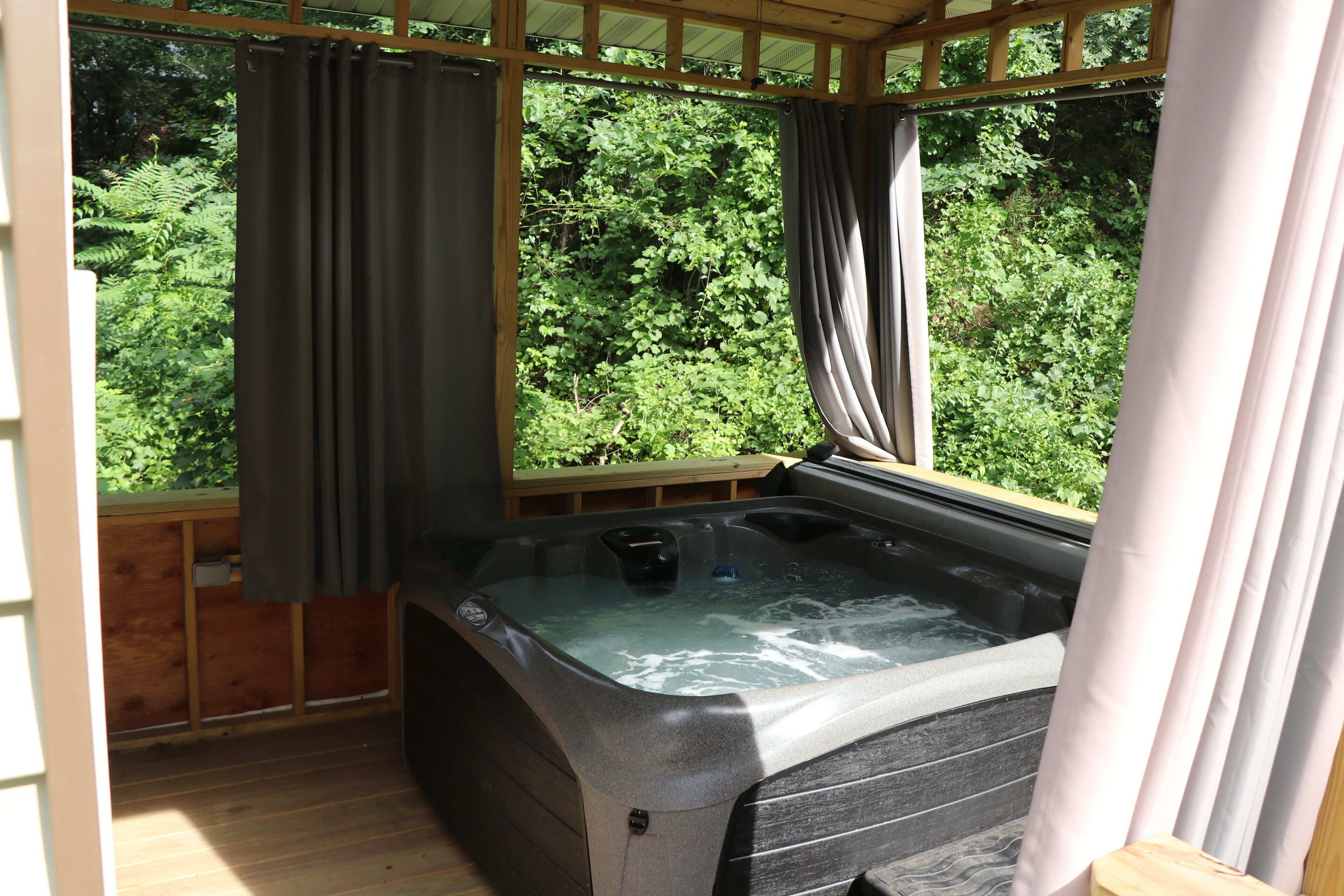 A hot tub in a wooden deck area with curtains, surrounded by lush greenery.