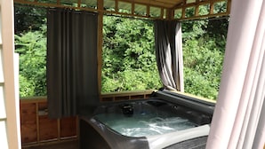 A hot tub in a wooden deck area with curtains, surrounded by lush greenery.