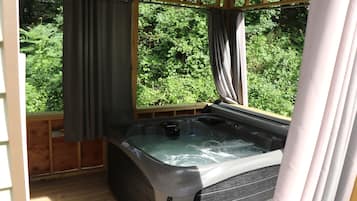 A hot tub in a wooden deck area with curtains, surrounded by lush greenery.
