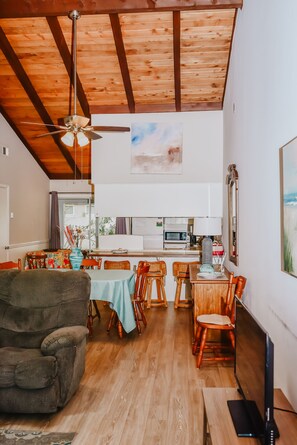 A spacious living area with a wooden ceiling, a dining table with chairs, a sofa, and a TV.