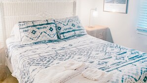Sängyssä on valkoinen ja sininen kuviollinen päiväpeite, valkoinen, kudottu pääty ja seinällä on kehystetty kuva.