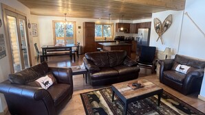 A spacious living room with leather sofas, a coffee table, and a rug. A kitchen area is visible in the background with wooden cabinets and a refrigerator. - Stewart Family Cabin by Face Lake (Logan Lake)