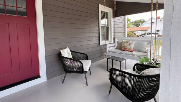 A covered porch with a red door, two black wicker chairs, a small table, and a white bench with cushions.