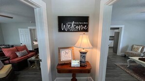 A hallway with a welcome sign, a lamp, and a framed picture on a wooden console table.