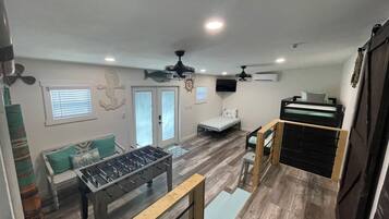 Un espacio habitable compacto con mesa de futbolín, cama, escritorio y ventilador de techo.