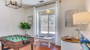 A room with a foosball table, a small table with a basket and magazines, a lamp, and a window with curtains. - Beautiful Hemet Home: Large Patio & Mtn Views! (Hemet)