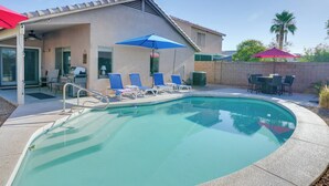 En swimmingpool med liggestole og parasoller, en overdækket terrasse og udsigt til palmer.