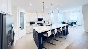 A modern kitchen with white cabinetry, a large island with bar stools, and a dining area with a round table and chairs. - Great location,Best value, beautiful home+games (Indio)