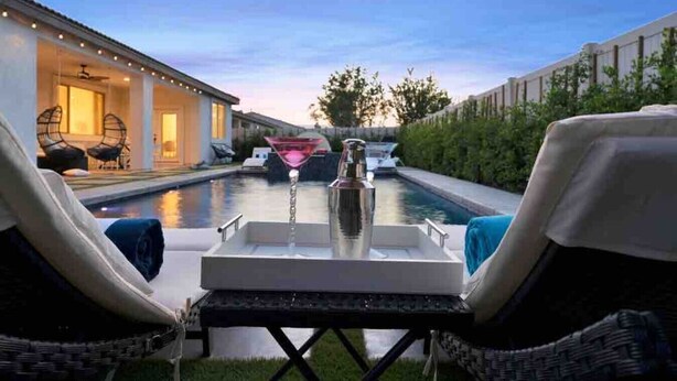 A poolside area with lounge chairs, a bar table with a martini glass, and a view of a building in the background. - Great location,Best value, beautiful home+games (Indio)