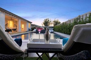 A poolside area with lounge chairs, a bar table with a martini glass, and a view of a building in the background. - Great location,Best value, beautiful home+games (Indio)