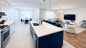 A modern kitchen with a central island, a dining area with a table and chairs, a living room with a sofa and TV, and a large window providing natural light. - Great location,Best value, beautiful home+games (Indio)