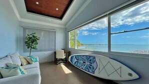 Una habitación con un ventanal grande que da a una playa, un sofá blanco con cojines estampados, una tabla de surf apoyada contra la pared y un techo con paneles de madera.