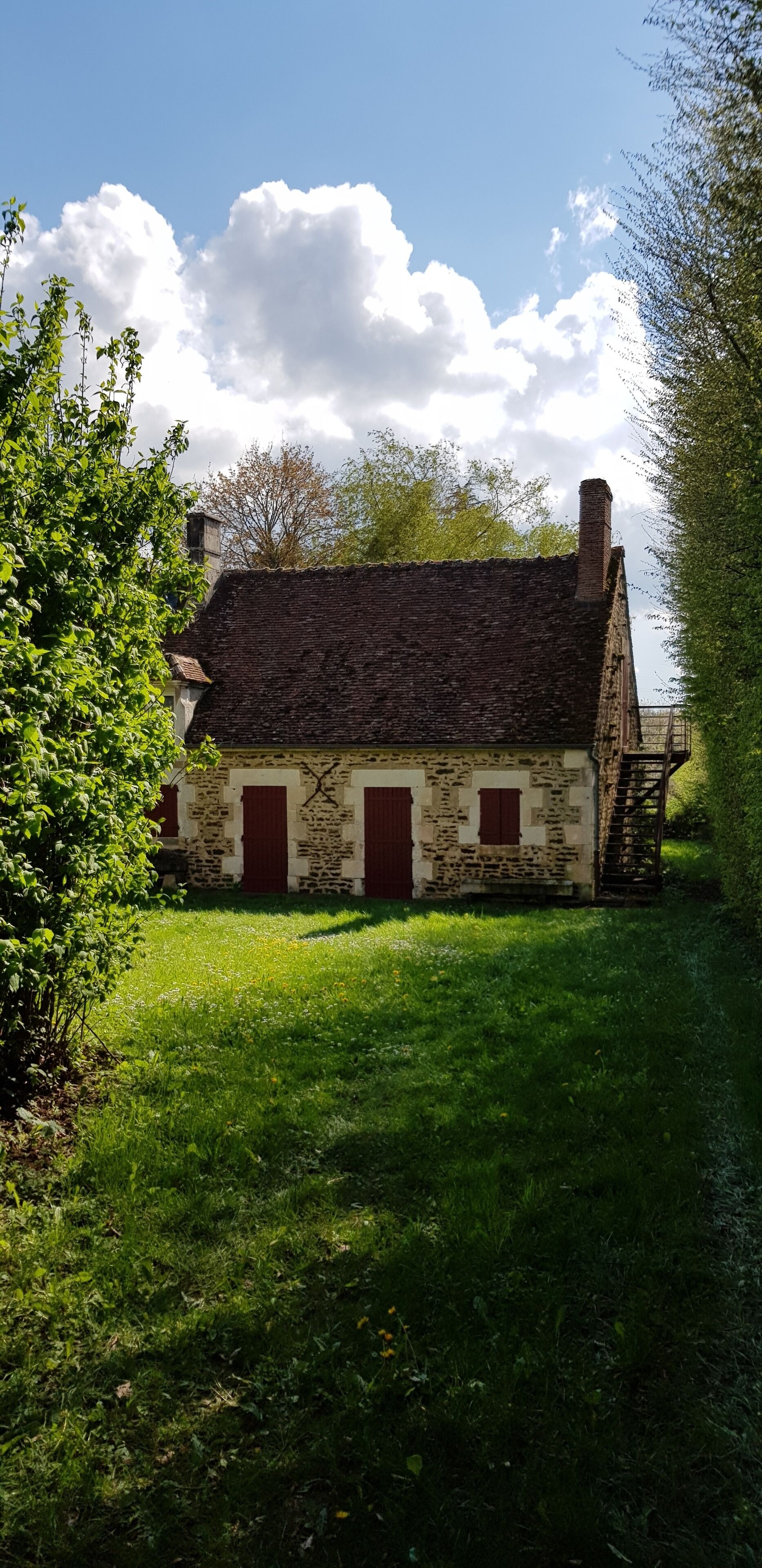 Een gebouw van steen met een schuin dak en rode luiken, omgeven door groen en een heldere lucht.