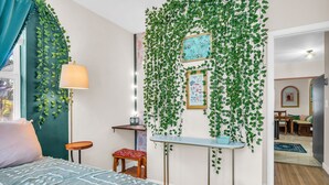 A bedroom with a bed, a bedside table, a lamp, a small stool, and a wall adorned with green plants and framed pictures.