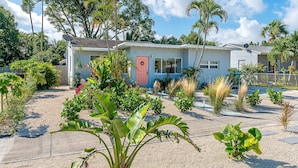 A single-story house with a red door, surrounded by a variety of tropical plants and a gravel driveway.