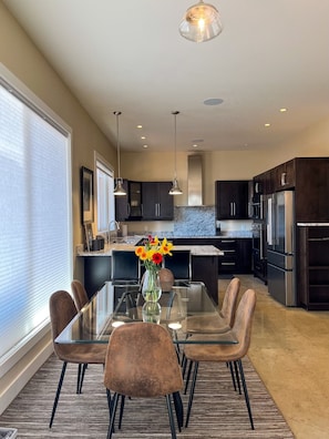 A modern kitchen with dark cabinets, stainless steel appliances, and a glass dining table with brown chairs.