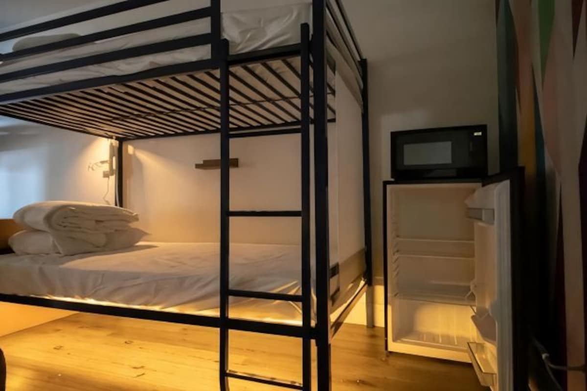 Uma cama beliche com televisão embutida, um pequeno frigorífico e chão de madeira.