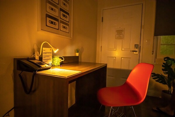 Un petit bureau avec une lampe, une chaise rouge, une porte et un mur orné de tableaux ou d’objets encadrés.