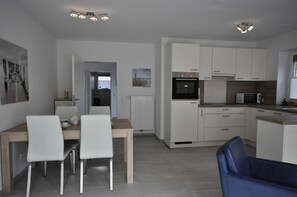 Una cocina moderna con armarios blancos, horno integrado y microondas. Hay una zona de comedor con una mesa de madera y dos sillas blancas.