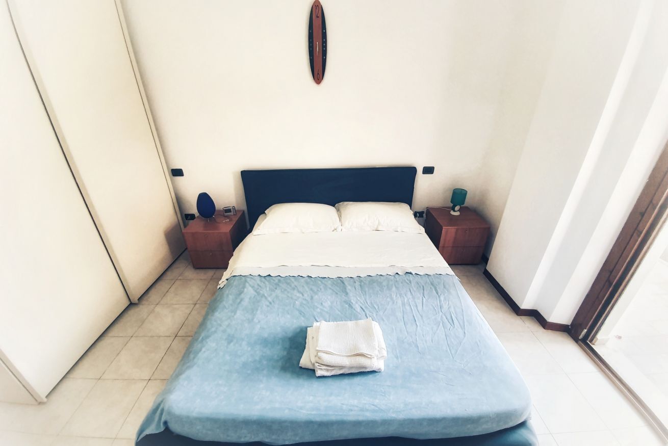 Una cama bien hecha con colcha azul y almohadas blancas, dos mesitas de noche con lámparas, y un reloj de pared.