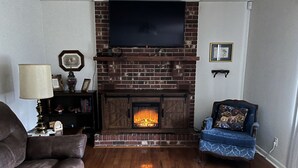 A living room with a brick fireplace, a television mounted above it, a blue armchair with a bear-themed pillow, and a leather sofa. - Mamaws mountain view Quiet country setting in the middle of a lot of attractions (Fletcher)