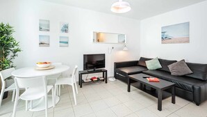 Una sala de estar moderna con un sofá de cuero negro, una mesa de comedor blanca y sillas. Un televisor de pantalla plana está montado en la pared, y hay varias fotografías enmarcadas.