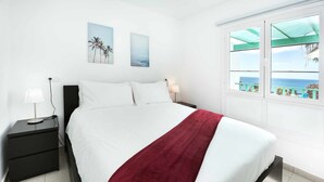 Una cama bien tendida con ropa de cama blanca y roja, dos mesitas de noche con lámparas y una ventana con vista al mar.