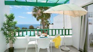 Un balcón con vista al mar, una mesa con sillas, una planta en maceta y una chaqueta amarilla colgada de una silla.