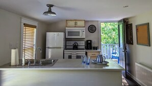 Eine kleine Küche mit Spüle, Mikrowelle, Wasserkocher und Kaffeemaschine. Ein Fenster mit Jalousien, eine Wanduhr und eine Tür nach draußen sind vorhanden.