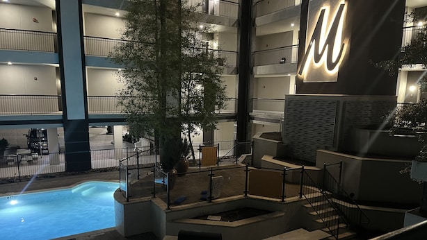 Un espace piscine intérieur moderne, doté d’un grand panneau lumineux en forme de « W », entouré de nombreux balcons et d’un escalier.