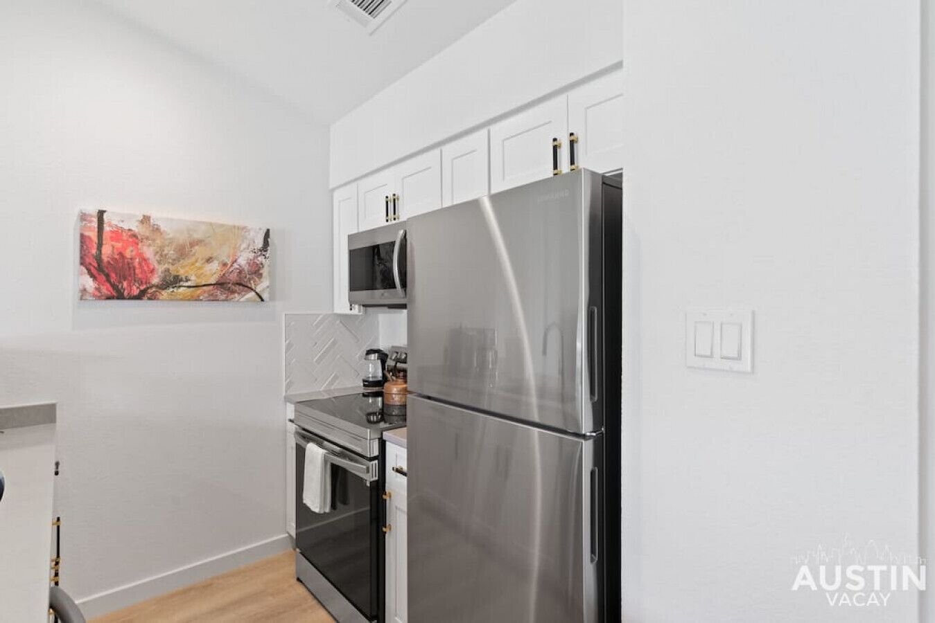 A compact kitchen with a stainless steel refrigerator, oven, and microwave. There is a small countertop with a kettle and a coffee maker. The kitchen has white cabinets and a herringbone-patterned backsplash.
