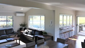 A spacious living area with a large window, a leather sofa, a dining table, and a bar area with stools. - Romantic lakehouse set in a private vineyard in the Hunter Valley (Lovedale)