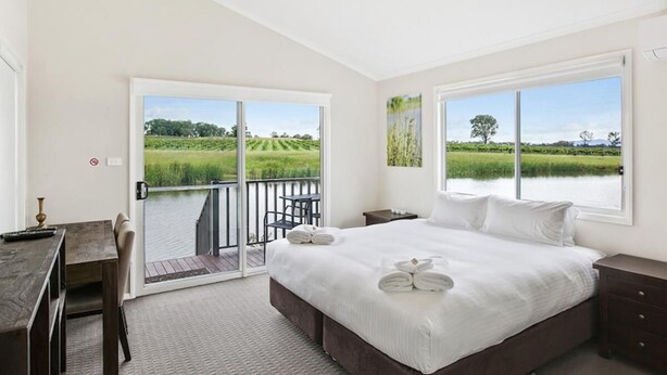 A bedroom with a large bed, a desk, and a balcony with a view of a green landscape. - Romantic lakehouse set in a private vineyard in the Hunter Valley (Lovedale)