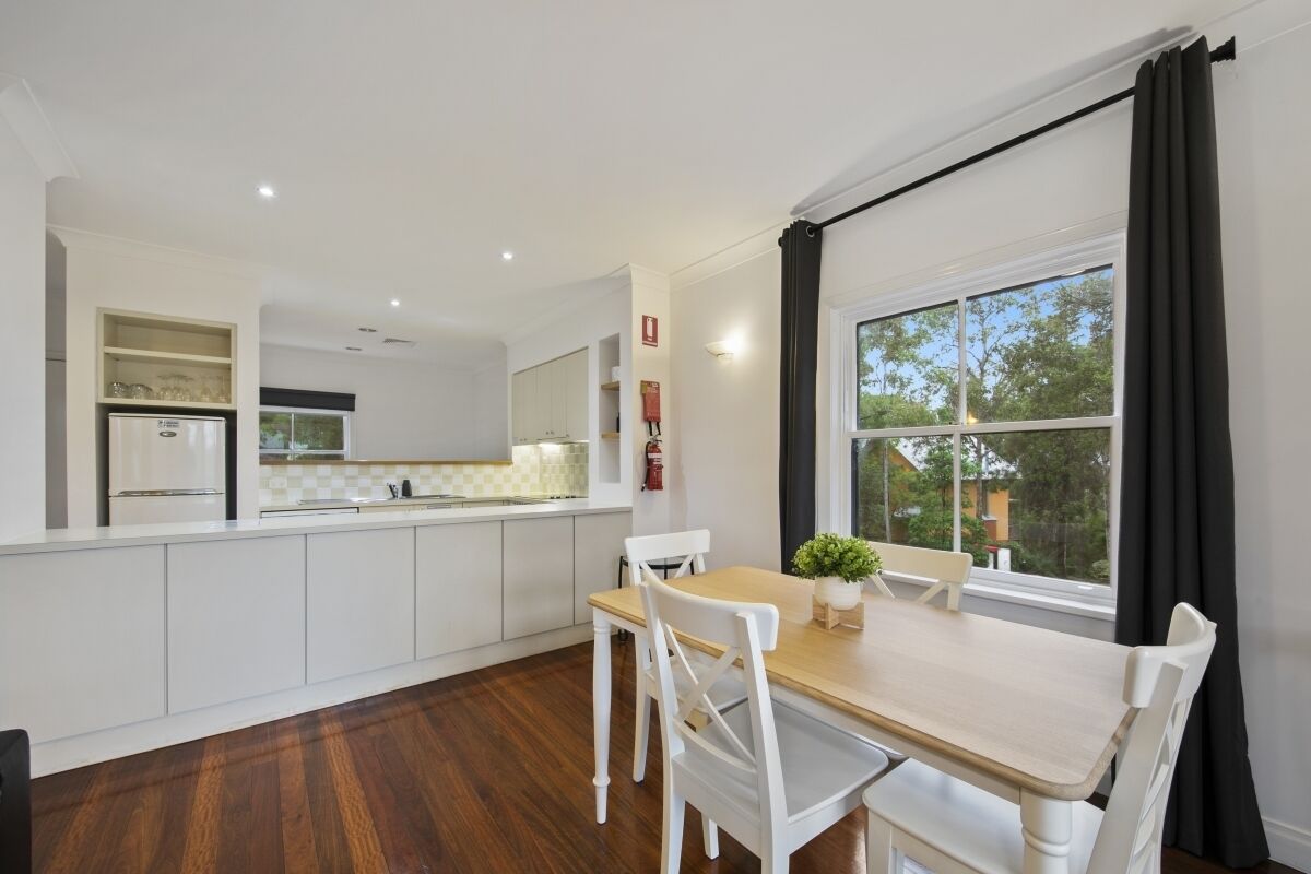 A modern kitchen with white cabinetry, a wooden dining table, and a large window with black curtains.