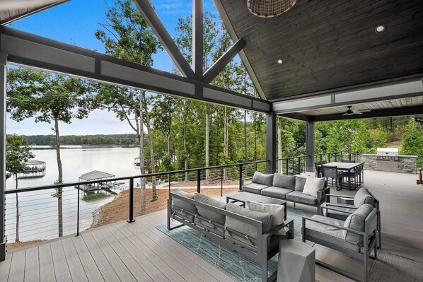 A covered outdoor deck with a sofa, chairs, and a grill, overlooking a lake with dock structures.