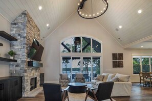 A spacious living room with a stone fireplace, large windows, a dining area, and a high ceiling with recessed lighting.