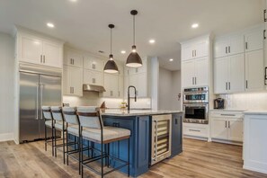 A modern kitchen with white cabinets, a stainless steel refrigerator, a double oven, and a bar area with stools.
