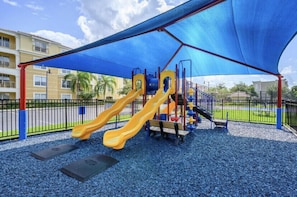 Ein Spielplatz mit zwei gelben Rutschen und einer Kletteranlage, die von einer blauen Markise überspannt ist, mit einem Zaun und Gebäuden im Hintergrund.