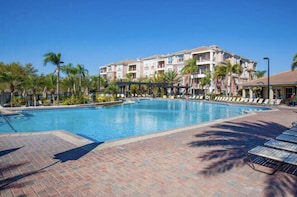 Ein Swimmingpool mit Liegestühlen und Palmen, umgeben von einer Ziegelterrasse und einem mehrstöckigen Gebäude mit Balkonen.