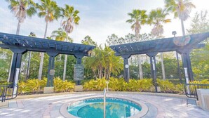 Ein runder Pool mit einer zentralen Stütze, umgeben von einer gepflasterten Fläche mit Palmen und einer Pergola.