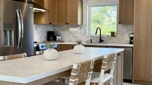 Ett modernt kök med träskåp, ett kylskåp i rostfritt stål, en inbyggd ugn, en stor köksö med barstolar och ett fönster med utsikt över grönområden.