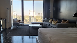 A modern hotel room with a large bed, a sofa, a chair, a coffee table, and a TV. The room has a city view through large windows.