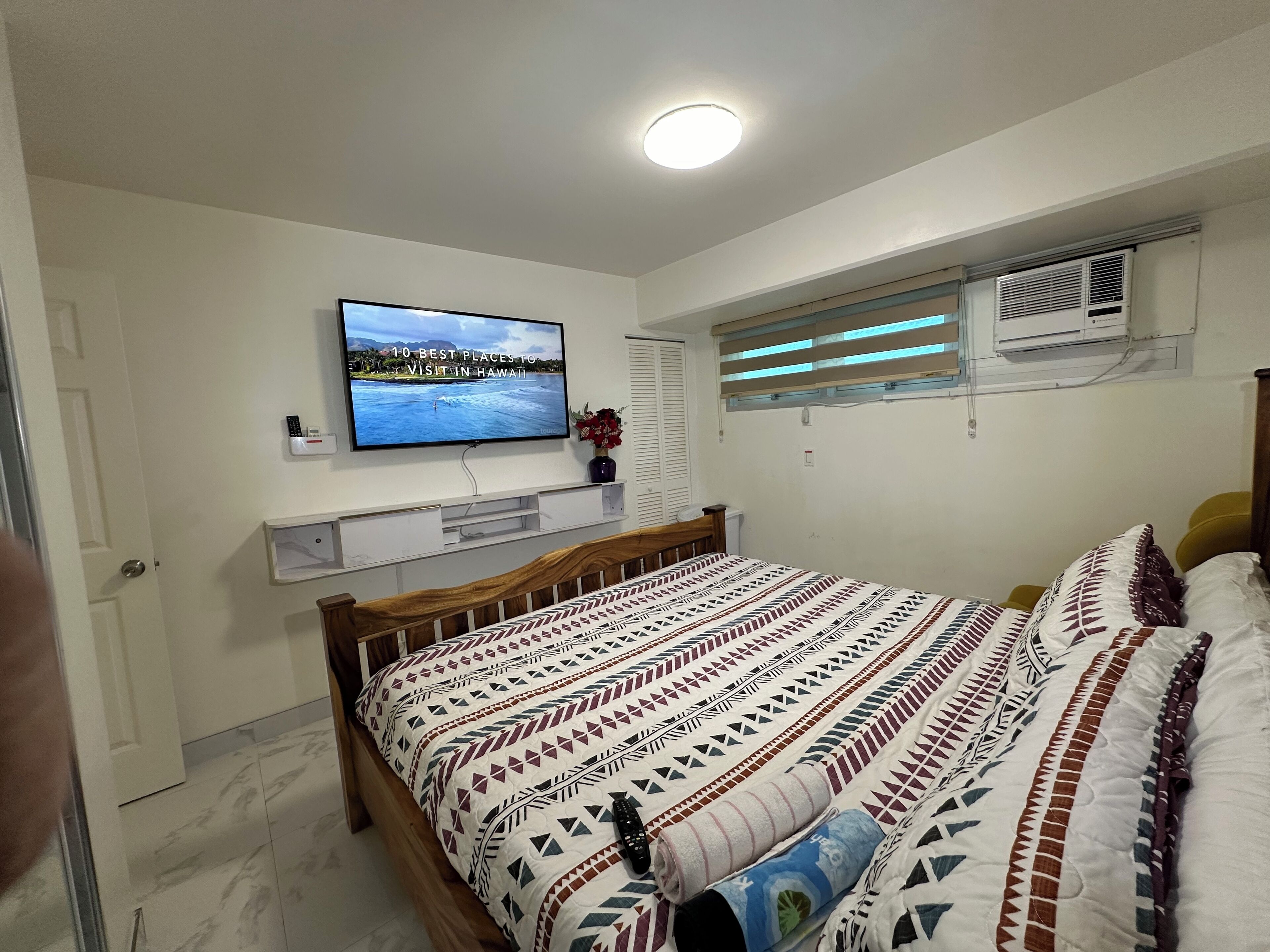 A bedroom with a bed, a television mounted on the wall, a window with blinds, and an air conditioning unit.