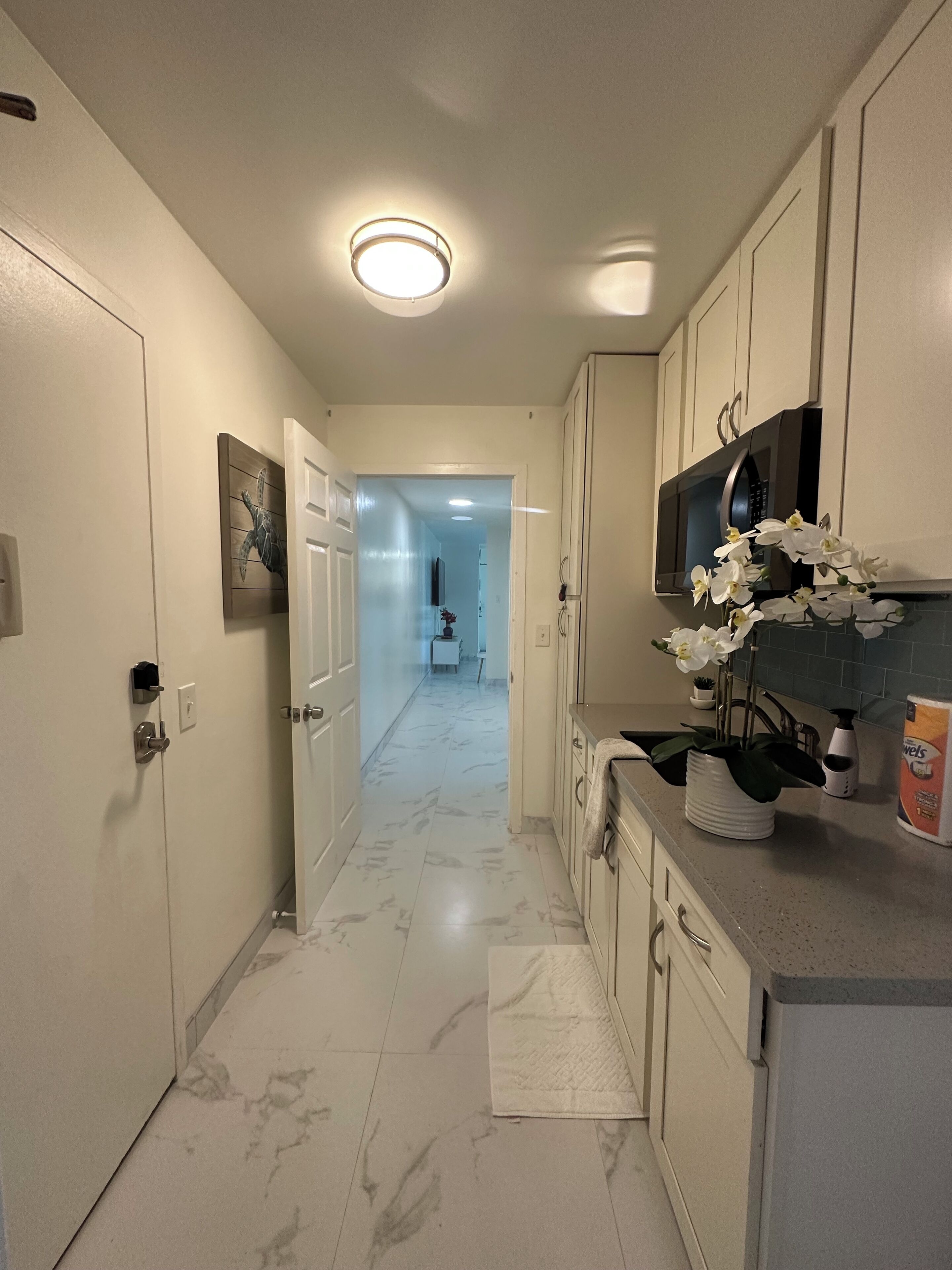 Un pasillo estrecho con suelo de mármol, paredes blancas y una lámpara en el techo. Hay una puerta a la izquierda y una zona de cocina a la derecha con armarios blancos y un microondas.