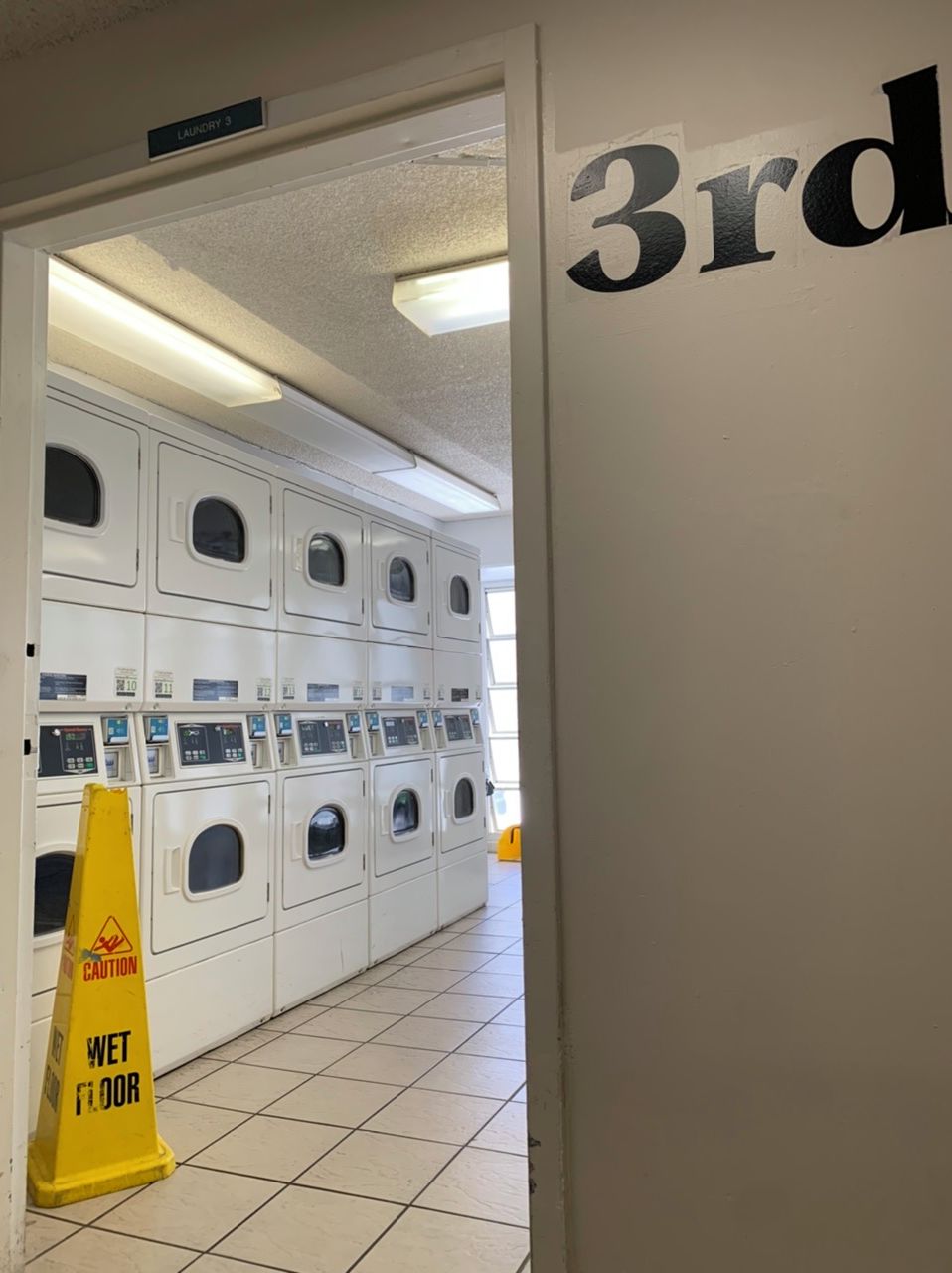 A laundry room with multiple washing machines, a caution sign on the floor, and a door labeled "3rd".