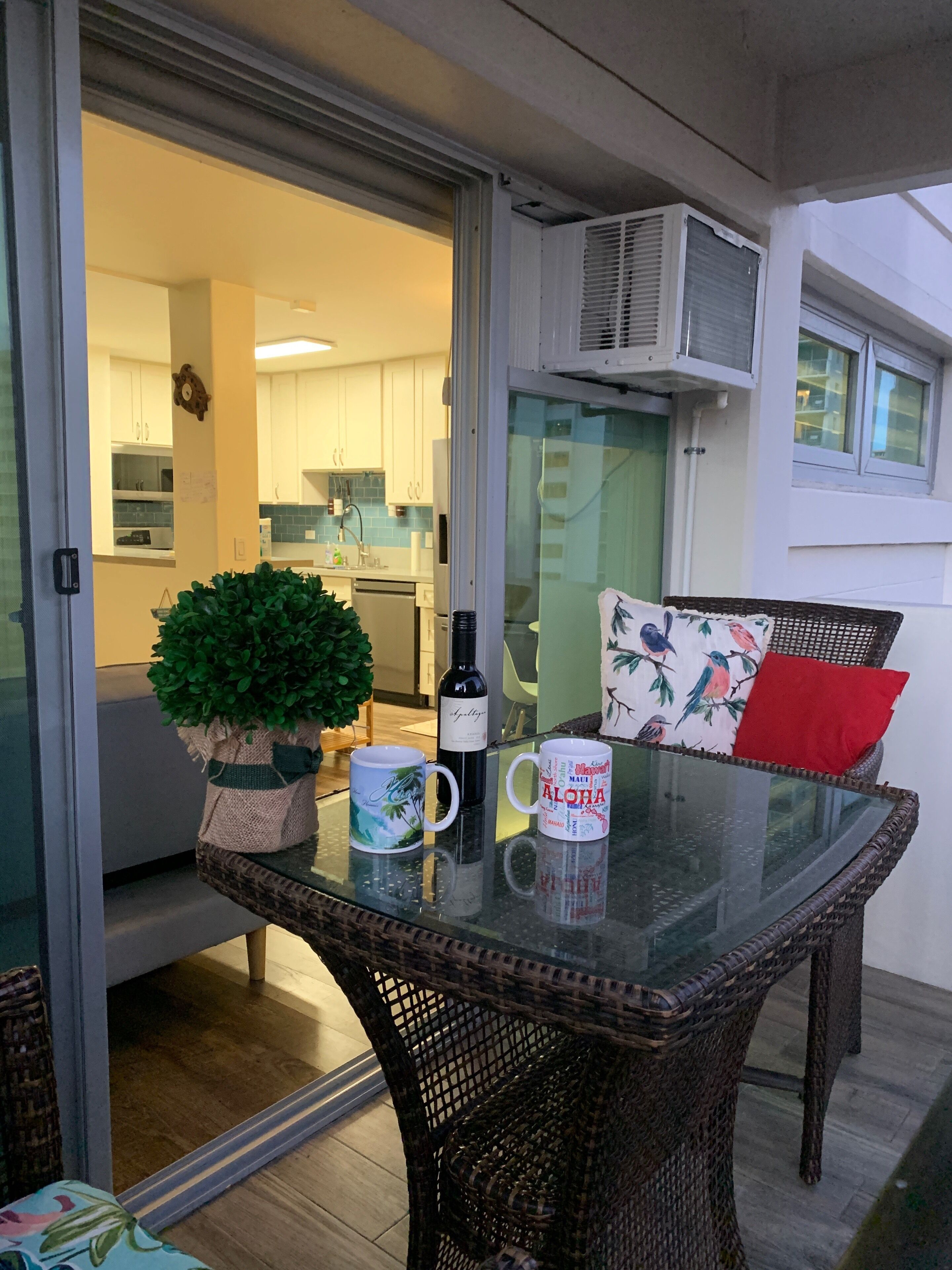 Une table à plateau en verre, ornée d’un pot de plante, de deux tasses et d’une bouteille, sur un balcon agrémenté d’une chaise en osier et de coussins.