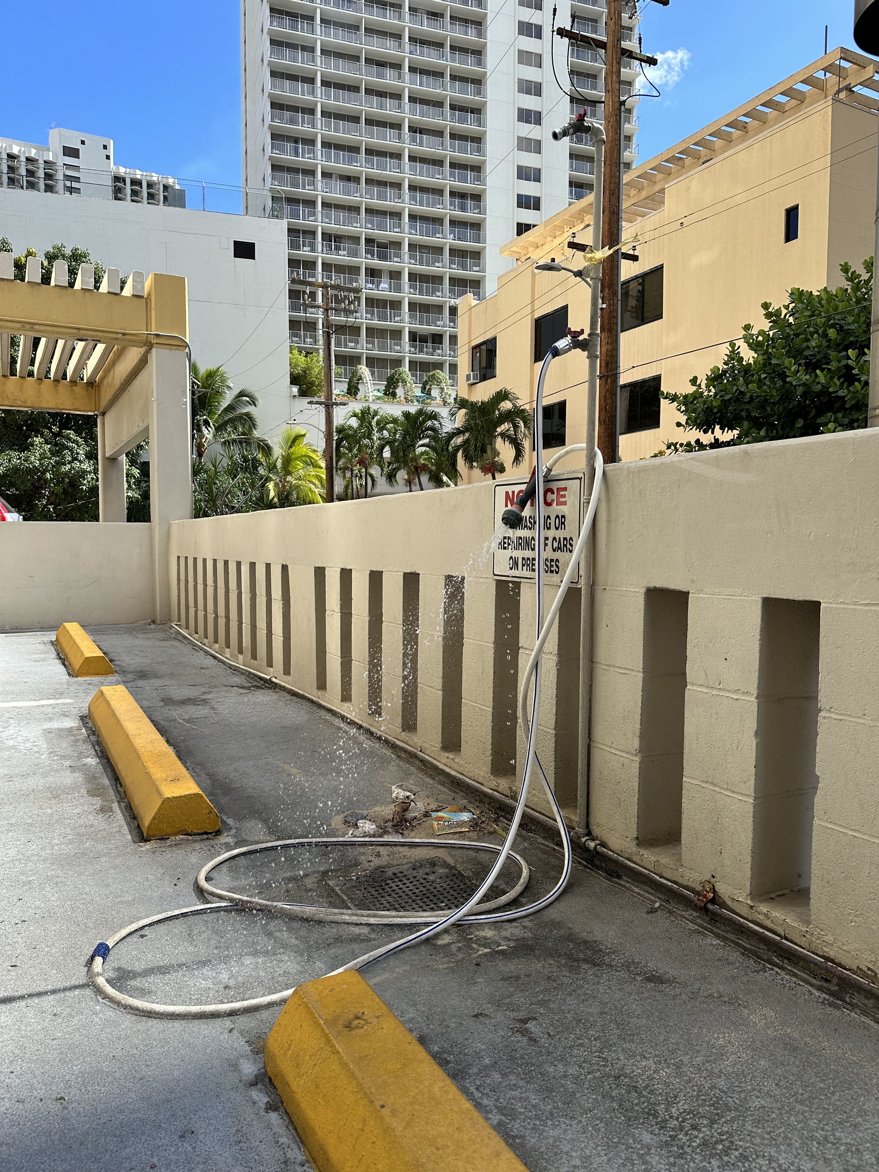 A parking area with a hose connected to a building, a 'No Washing or Repairing of Cars' sign, and a yellow bollard.