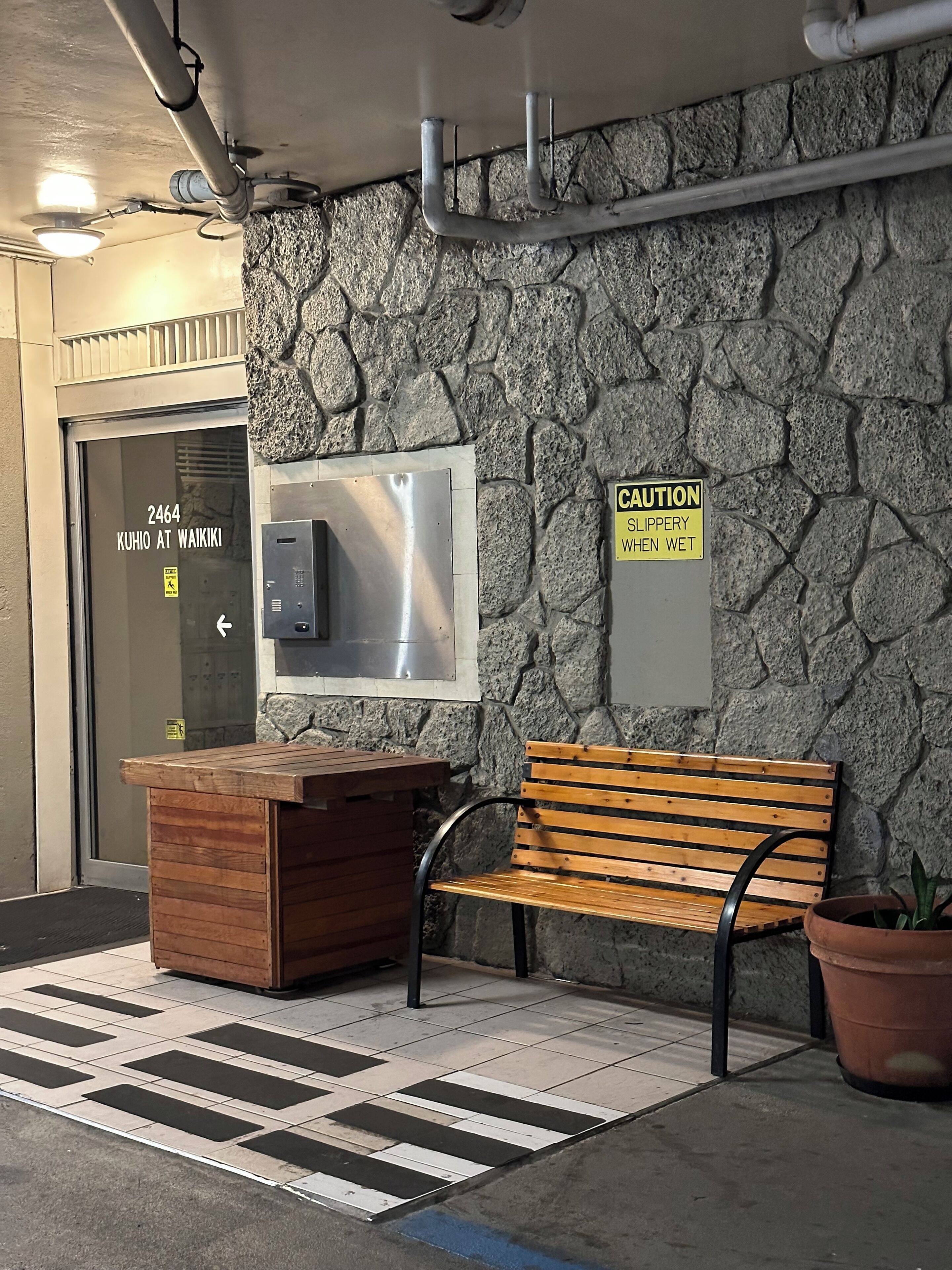 A stone wall with a caution sign, a bench, a trash bin, and a door labeled "2464 KUHIO AT WAIKIKI".