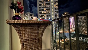 Een balkon met een rieten tafeltje, een bloevas en een uitzicht op een stadsbeeld 's nachts.
